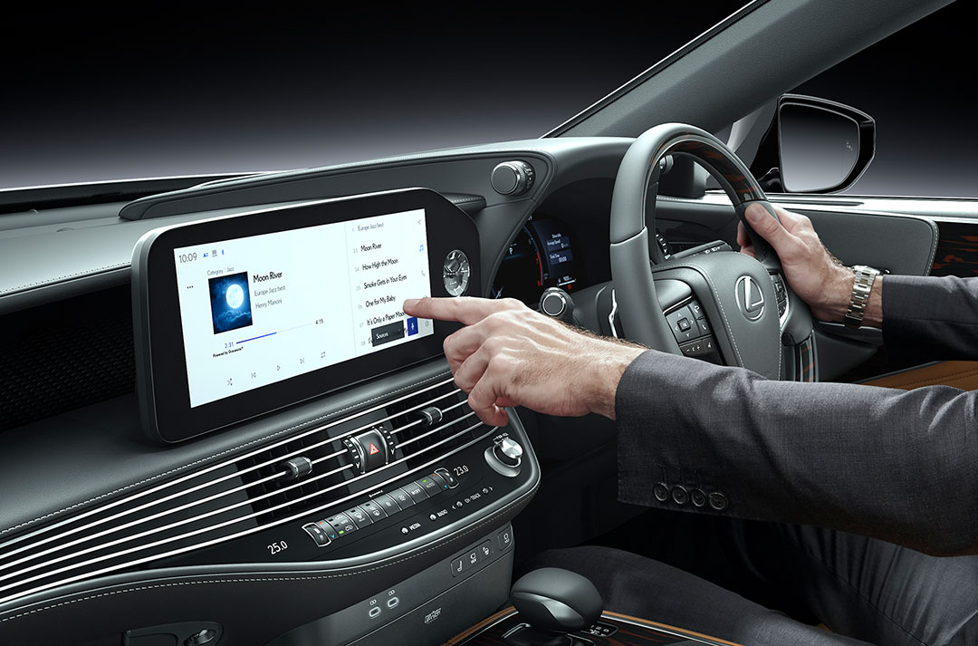 The driver's left hand reaches to the multimedia screen of the Lexus LS.