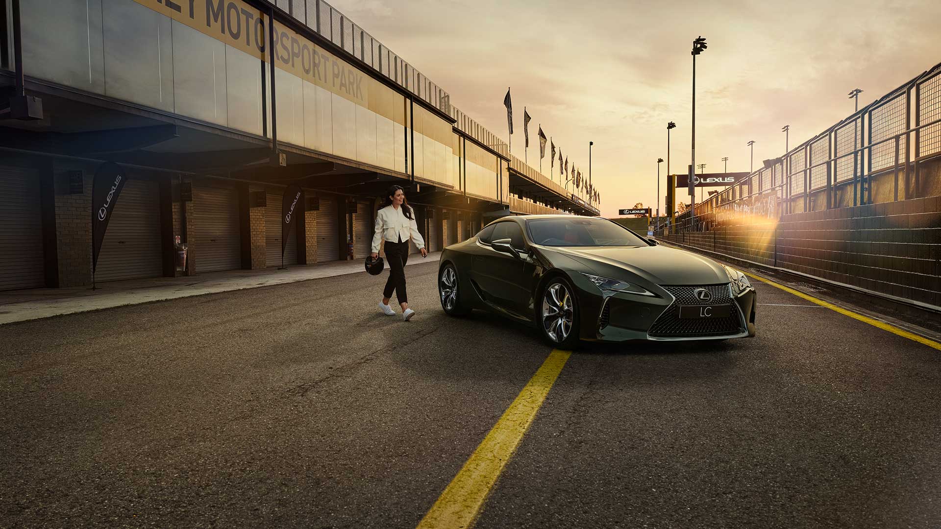 A woman dressed in a white jacket and black pants and holding a helmet walks towards an LC coupe parked in a pit lane